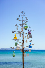 A coastal art installation features a tree adorned with colorful decorations, set against a bright...