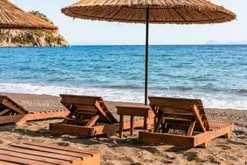 Wooden loungers with straw umbrellas line the sandy beach while gentle waves lap at the shore under a clear blue sky