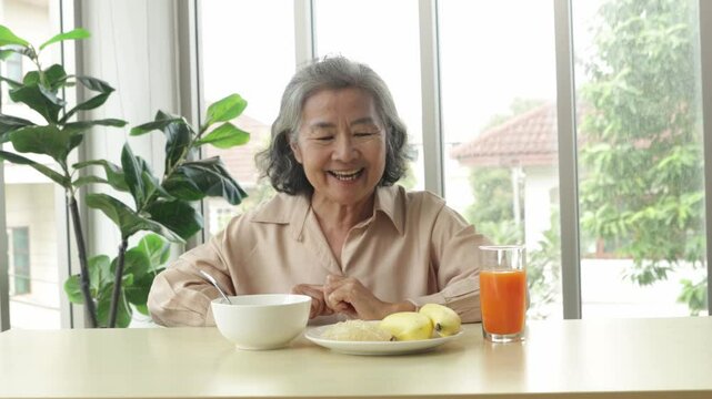 Healthy elderly Asian woman sits down to eat lunch with meatless, easily digestible congee and a banana and freshly squeezed orange juice rich in vitamin C with satisfaction.