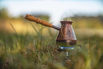 Aromatic coffee steams in copper engraved coffee pot on gas burner with blue flame among grass....