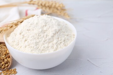 Wheat flour in bowl, grains and spikes on white table, closeup. Space for text