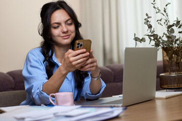 beautiful woman in blue shirt holds smart phone uses laptop in home office