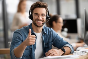 Young man with headset smiling and giving thumbs up while working at a desk in a modern office environment, showcasing customer service and teamwork in action