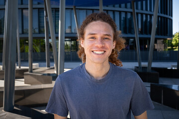 Smiling young man with curly hair and earring standing outdoors in modern urban environment. Casual style, sunny day, contemporary lifestyle concept.