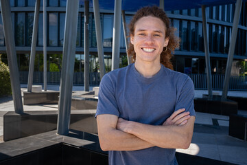 Confident young man with curly hair smiling with arms crossed in modern urban setting. Casual fashion, contemporary lifestyle, positive attitude concept.