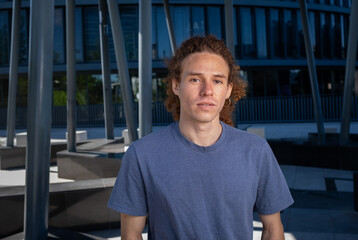 Serious young man with curly hair and earring standing in modern urban environment. Casual fashion, contemporary lifestyle, confident and neutral male portrait.