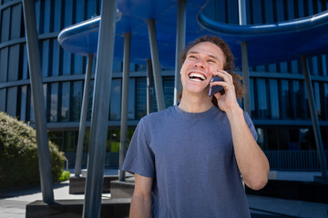 Young man with curly hair laughing and talking on smartphone outdoors, joyful communication and modern lifestyle in urban city environment