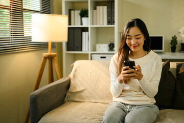 Obraz premium Young Asian Woman Relaxing at Home with Smartphone on Cozy Sofa
