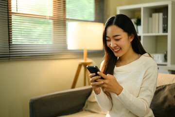 Obraz premium Close-up image of a smiling young Asian woman using her smartphone, Bright modern home interior with warm lighting, and a calm