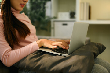 Fototapeta premium Close-up of a woman typing on a laptop while sitting comfortably on a sofa with a cushion on her lap, Home Based Digital Work