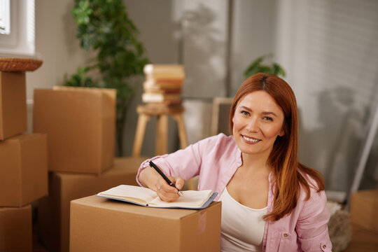 A woman sits on a cardboard box in a room filled with moving boxes, writing in a notebook to plan her upcoming relocation and organize her new home.