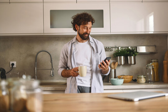 A man stands in a contemporary kitchen, sipping coffee from a mug, while looking at his smartphone with a content smile during a relaxed morning.