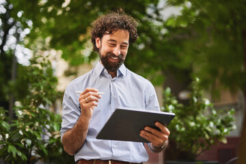 A man with curly hair and a beard is smiling as he uses a tablet in a green, bustling outdoor...
