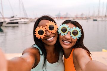 Two happy multiracial female friends wearing flower-shaped sunglasses taking a selfie by the sea
