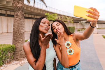 Two multiracial female friends blowing kisses at the camera while taking a selfie with a smartphone