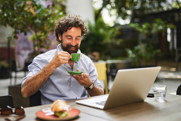 A man sits at an outdoor table, smiling as he enjoys a cup of coffee and a light snack while...