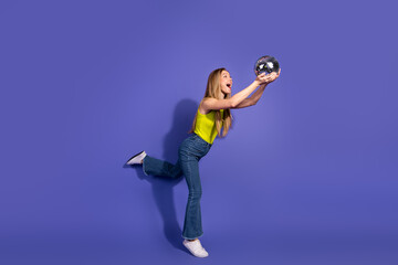 Cheerful young woman in a stylish yellow top holding a disco ball, standing on one leg against a...