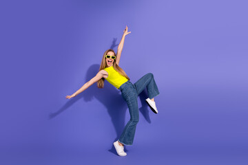 Joyful young woman dancing with enthusiasm against a bright purple background wearing a stylish yellow top and sunglasses