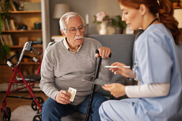 A red-haired nurse in scrubs sits next to a mature man in a grey sweater, reviewing his medication...