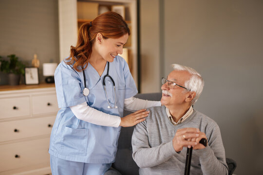 In a bright living room, a smiling, female home nurse in scrubs gently places her hand on the shoulder of an elderly gentleman. He sits holding a cane, looking up at her.