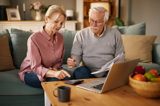 An elderly Caucasian couple is sitting on their couch, reviewing documents and using a laptop and calculator. They are planning their finances and looking over their life insurance policy.