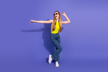 Joyful young woman dancing in casual outfit with a vibrant yellow top against a purple background,...
