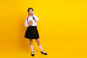 Cheerful schoolgirl in uniform holding a phone and expressing surprise on a yellow background,...