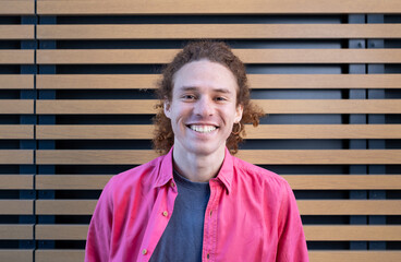 Smiling young man with curly hair wearing pink jacket standing against modern wooden wall. Casual fashion, cheerful male portrait, contemporary lifestyle concept.