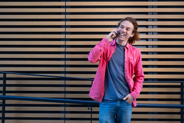 Young man with curly hair in pink shirt smiling and talking on smartphone outdoors against modern wooden wall. Casual student, technology and youthful lifestyle concept.