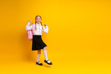 Happy schoolgirl posing in uniform with backpack on vibrant yellow background representing...