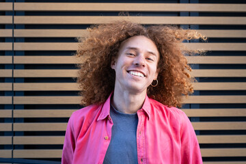 Smiling young man with curly hair wearing pink jacket and t-shirt against modern wooden wall. Youthful fashion, student lifestyle, personal branding and future entrepreneur concept.