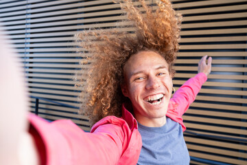 Cheerful young man with curly hair taking dynamic selfie outdoors in pink jacket. Joyful student, social media lifestyle, self expression and youthful energy concept.