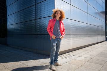 Young man with curly hair and pink jacket standing outdoors in front of modern metallic building, urban fashion and student lifestyle portrait, future entrepreneur and contemporary business concept