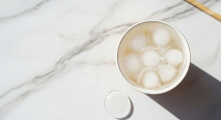 A top view of a white cup filled with round ice balls on a marble surface. A small lid is placed beside the cup, creating a clean and minimalistic scene.