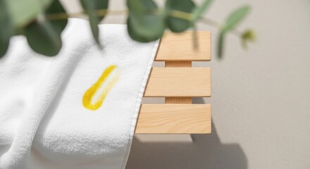 A close-up of a wooden bench with a white towel draped over it. A small yellow oil drop is visible on the towel. Green eucalyptus leaves are in the background.