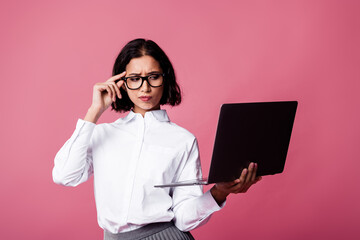 Charming businesswoman in glasses holding a laptop and showcasing a thoughtful gesture against a vibrant pink background