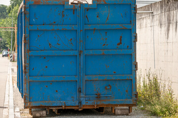 Blue container with steel rolls for waste materials is standing at the side of the road, close to a concrete wall.