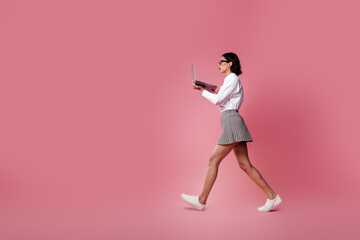 Fashionable young woman walking confidently while working on a laptop against a vibrant pink background