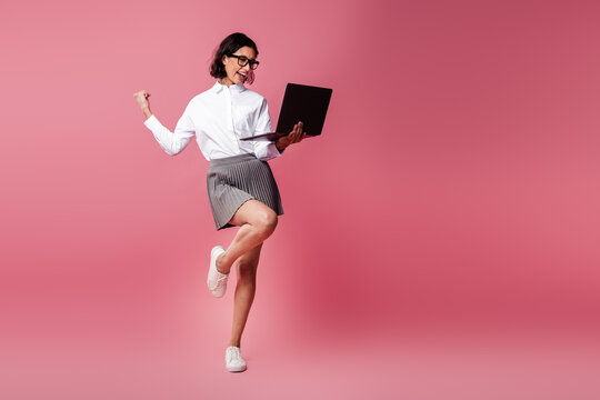 Charming young businesswoman in formal attire celebrating success with a laptop against a vibrant pink background - Powered by Adobe