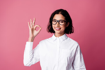 Confident young woman in glasses giving an okay hand gesture on a bright pink background in formal...