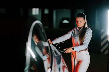 Woman charges electric car in neon light at urban charging station during nighttime hours