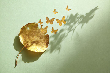 Single golden leaf with delicate butterfly shapes emerging from it, casting soft shadows on pale green surface, evoking sense of transformation and lightness
