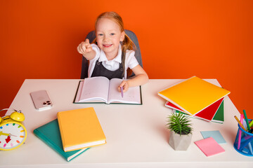Cheerful schoolgirl studying at a desk with colorful school supplies under bright orange...