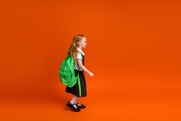 Adorable young schoolgirl with green backpack walking against bright orange background showcasing primary school theme