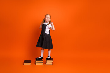 Adorable young girl standing confidently on books against vibrant orange background embracing academic spirit and joy of learning
