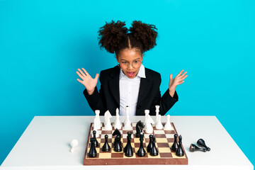 Charming schoolgirl playing chess enthusiastically against a turquoise background, exhibiting...