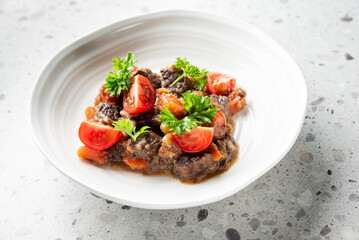 Beef stew served on a white plate, garnished with parsley and cherry tomato halves, presented on a speckled countertop in a gourmet culinary setting.