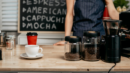 A young Asian female barista in a striped apron carefully pours steamed milk to create intricate latte art, working in a cozy modern caf&eacute; with professional coffee equipment and takeaway cups.