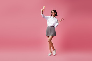 Young stylish woman taking a selfie on a pink background, wearing trendy formal attire and a pleated skirt