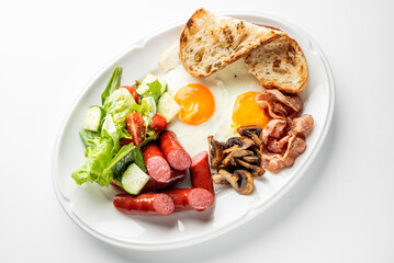 Traditional English breakfast with sunny-side-up eggs, bacon, sausages, toast, mushrooms, and fresh salad on a white plate, shot from above on a table.